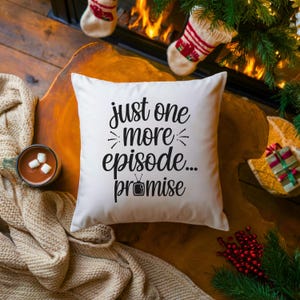 May include: A white square pillow with the text "just one more episode... promise" in black script. The pillow is on a wooden surface, with a cup of hot chocolate and Christmas decorations in the background.