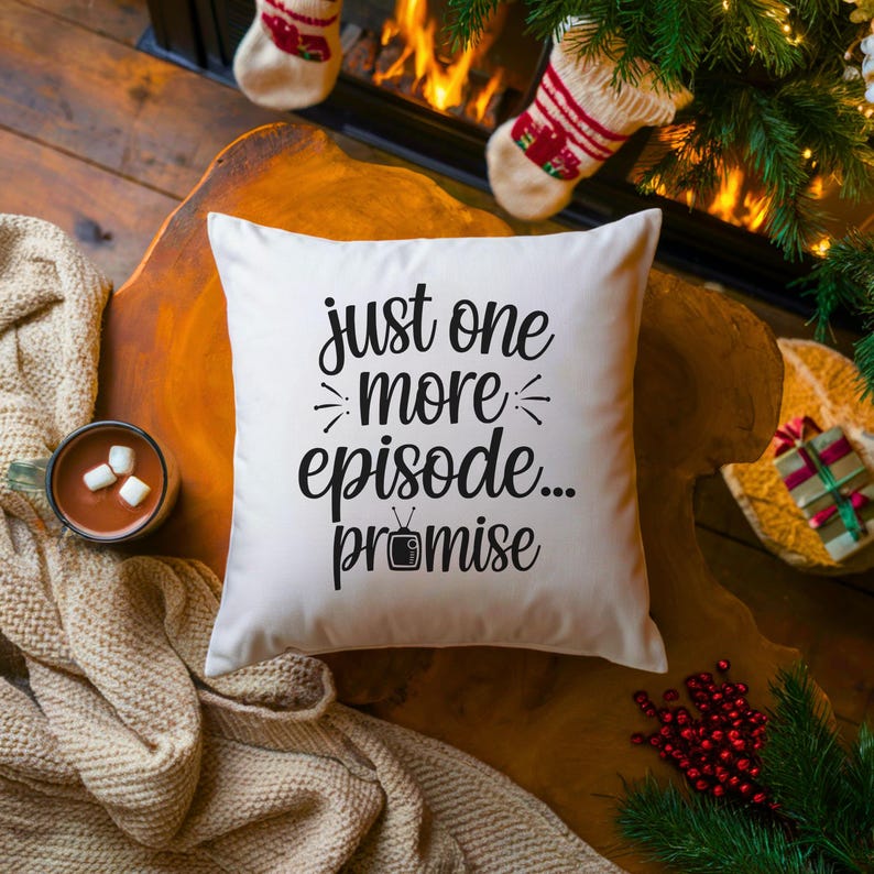 May include: A white square pillow with the text "just one more episode... promise" in black script. The pillow is on a wooden surface, with a cup of hot chocolate and Christmas decorations in the background.