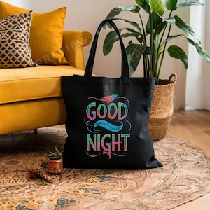 May include: A black tote bag with the words "GOOD NIGHT" in colorful, stylized lettering. The design includes a feather and flowing lines. The bag has two black handles and is sitting on a patterned rug.