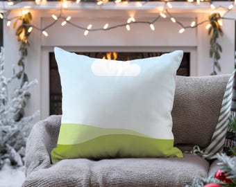 May include: A square decorative pillow with a landscape design. The pillow features a gradient sky blue background with a white cloud shape and green rolling hills at the bottom. The pillow is placed on a gray sofa.