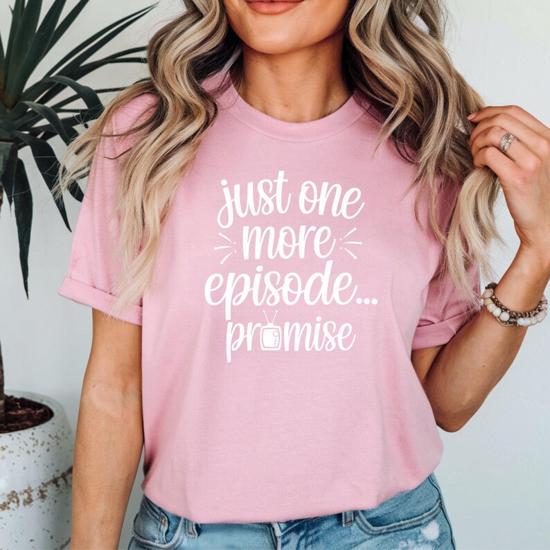 May include: A light pink t-shirt with the text "just one more episode... promise" in white script. The shirt features a TV graphic below the text. The short-sleeved tee is made of a soft material.