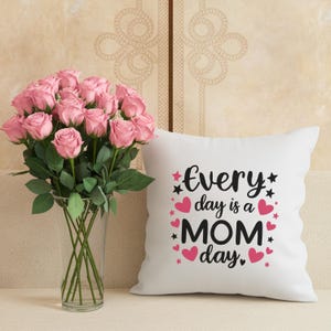 May include: A bouquet of pink roses in a clear glass vase next to a white pillow. The pillow has the text "Every day is a MOM day" in black script with pink hearts and stars.