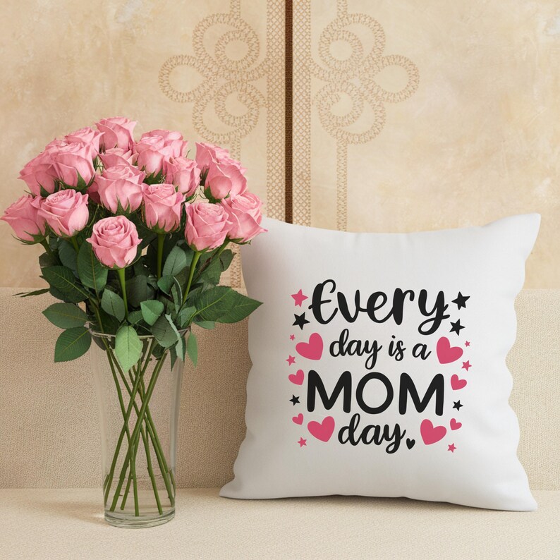 May include: A bouquet of pink roses in a clear glass vase next to a white pillow. The pillow has the text "Every day is a MOM day" in black script with pink hearts and stars.