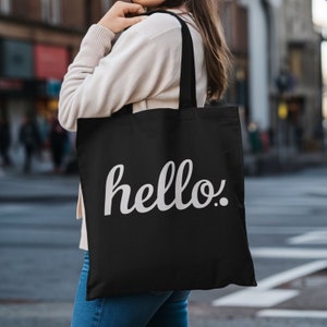 May include: Black tote bag with the word "hello." in white script. The bag has black handles and is made of a sturdy fabric. The bag is being carried on a city street.