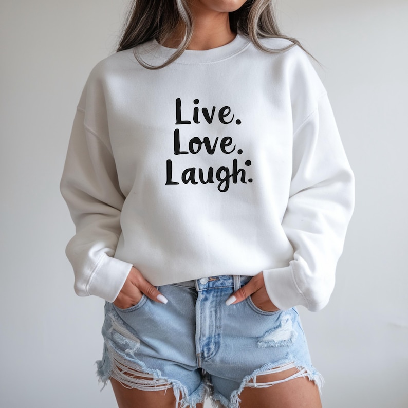May include: A white crew neck sweatshirt with the words "Live. Love. Laugh." printed in black. The sweatshirt is worn with blue denim shorts. The person is standing with their hands in their pockets.