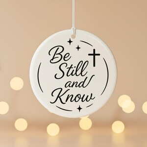 May include: A white, round ceramic ornament with the words "Be Still and Know" in black script. A black cross and star accents are also featured. The ornament hangs from a white string against a blurred background.