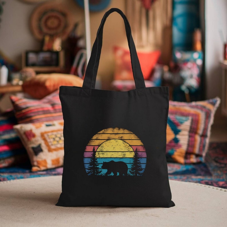 May include: Black tote bag featuring a sunset design with a bear silhouette, trees, and a colorful striped background. The bag has long handles for carrying over the shoulder. The design evokes a nature or outdoor theme.