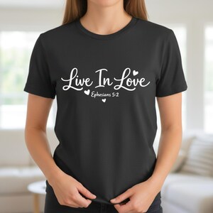 K&ouml;nnte beinhalten: Schwarzes T-Shirt mit dem wei&szlig;en Schriftzug "Live In Love" in Schreibschrift, verziert mit zwei kleinen Herzen. Darunter steht "Epheser 5:2". Das T-Shirt hat kurze &Auml;rmel.