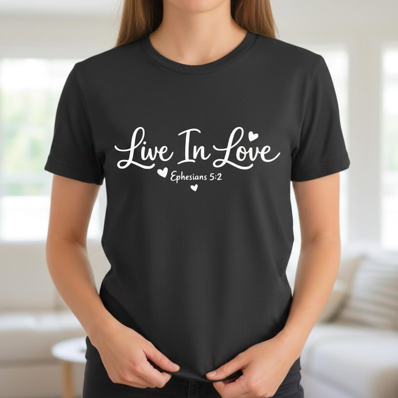 K&ouml;nnte beinhalten: Schwarzes T-Shirt mit dem wei&szlig;en Schriftzug "Live In Love" in Schreibschrift, verziert mit zwei kleinen Herzen. Darunter steht "Epheser 5:2". Das T-Shirt hat kurze &Auml;rmel.
