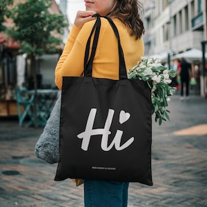 May include: Black tote bag with the word "Hi" in white script and a heart symbol. The bag also has additional text at the bottom. The bag is carried over a person's shoulder and contains a bouquet of white flowers.