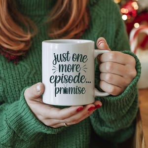 May include: White ceramic mug with the text "just one more episode... promise" in black script. The mug has a handle and is held by a person wearing a green sweater. The mug is a perfect gift for TV show lovers.