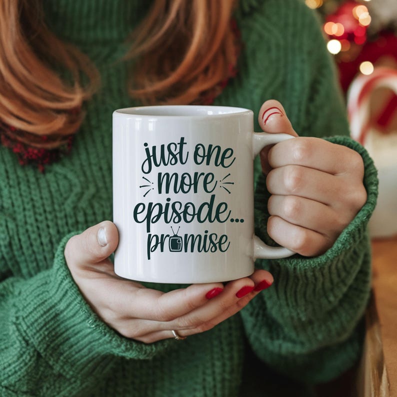 May include: White ceramic mug with the text "just one more episode... promise" in black script. The mug has a handle and is held by a person wearing a green sweater. The mug is a perfect gift for TV show lovers.