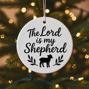 May include: A white, round ceramic ornament with the text "The Lord is my Shepherd" in black script. A black lamb silhouette and leafy sprigs are below the text. The ornament hangs from a silver string, set against a blurred background.