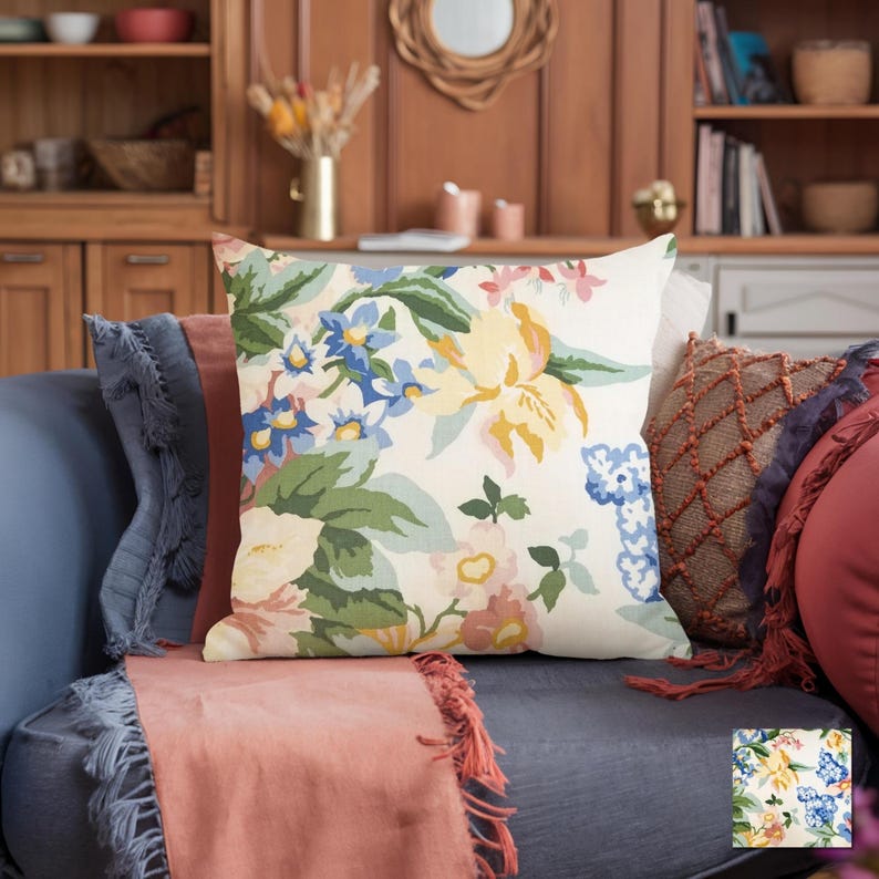 May include: A square decorative pillow with a floral pattern in yellow, blue, and pink on an off-white background. The pillow is on a blue sofa with other pillows and a coral-colored throw. The room has wooden furniture and decor.