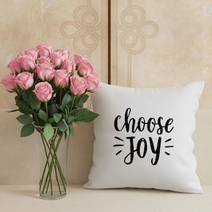 May include: A vase of pink roses stands beside a white cushion with the words "choose JOY" in black script. The roses are in a clear glass vase.