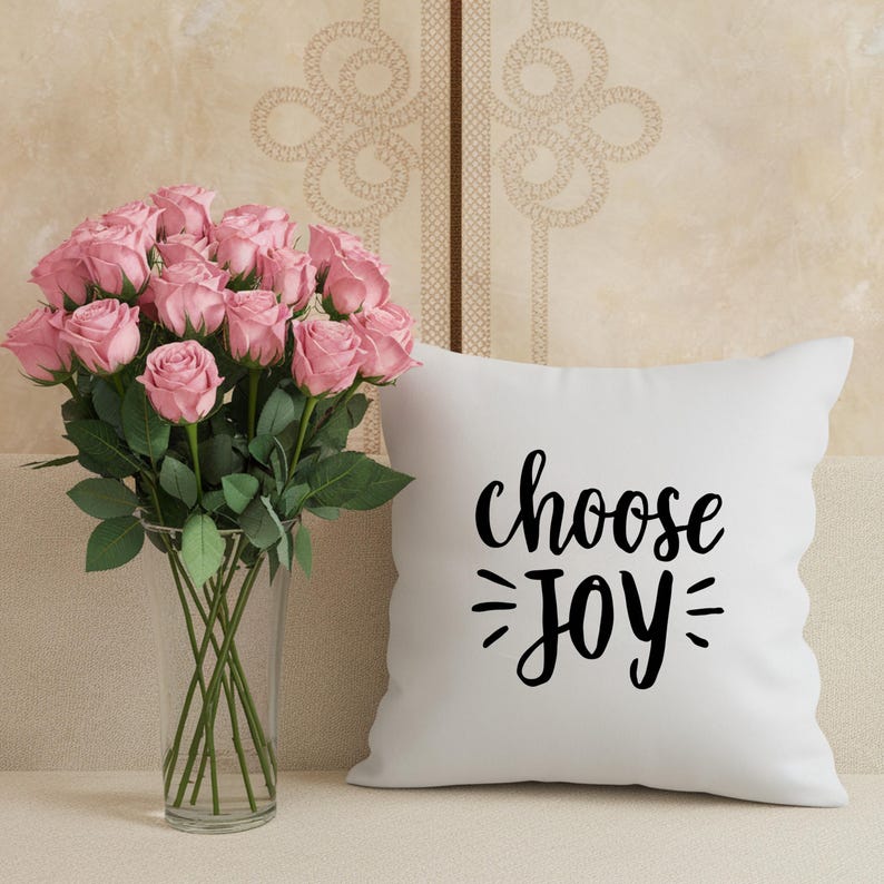 May include: A vase of pink roses stands beside a white cushion with the words "choose JOY" in black script. The roses are in a clear glass vase.