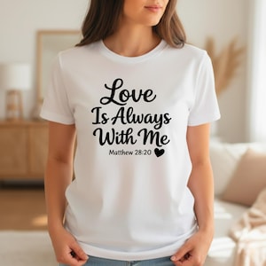 K&ouml;nnte beinhalten: Wei&szlig;es T-Shirt mit dem Text "Love Is Always With Me" in schwarzer Schrift. Darunter steht "Matthew 28:20" und ein schwarzes Herz. Das Shirt hat kurze &Auml;rmel und einen Rundhalsausschnitt.