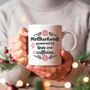 May include: White ceramic mug with the text "Motherhood: powered by love and caffeine." The text is in black with pink accents and floral designs. The mug is held by a person.