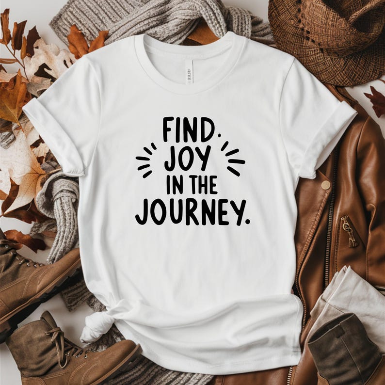May include: White t-shirt with the text "FIND JOY IN THE JOURNEY" in black. The shirt is displayed with a brown leather jacket, boots, and a hat, suggesting an autumn theme. The text is centered on the shirt.