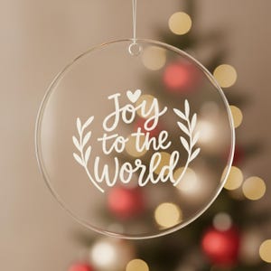 May include: Clear, round Christmas ornament with the words "Joy to the World" in white script. The ornament has a small loop at the top for hanging. The background is a blurred Christmas tree with red and gold ornaments.