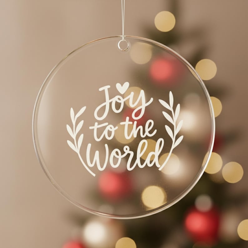 May include: Clear, round Christmas ornament with the words "Joy to the World" in white script. The ornament has a small loop at the top for hanging. The background is a blurred Christmas tree with red and gold ornaments.