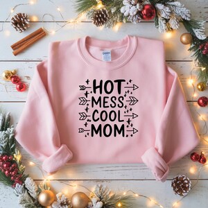 May include: A light pink sweatshirt with the text "HOT MESS COOL MOM" in black. The text is arranged in three lines with arrows and star accents. The sweatshirt has a crew neck and long sleeves.