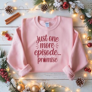 May include: A light pink sweatshirt with the text "just one more episode... promise" in burgundy script. The sweatshirt is a crew neck style and is folded on a white wooden surface with holiday decorations.