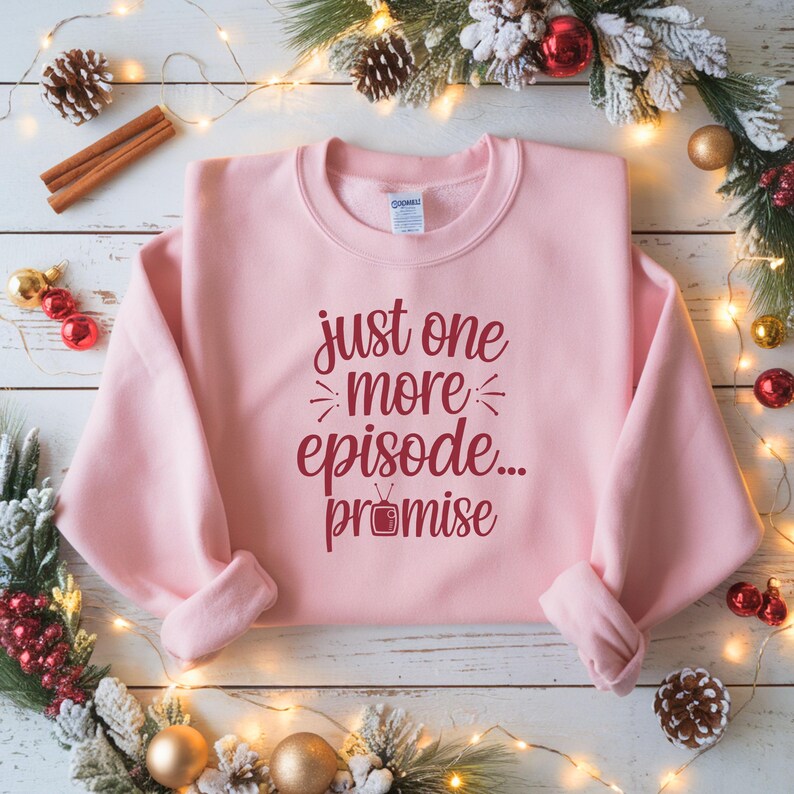 May include: A light pink sweatshirt with the text "just one more episode... promise" in burgundy script. The sweatshirt is a crew neck style and is folded on a white wooden surface with holiday decorations.