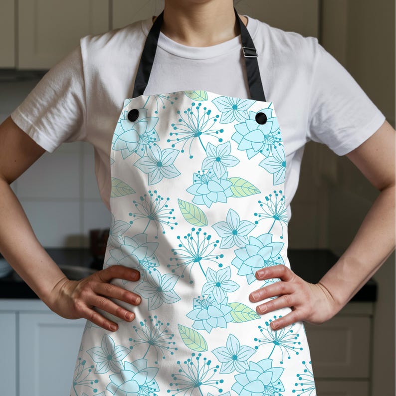 K&ouml;nnte beinhalten: Wei&szlig;er K&uuml;chensch&uuml;rze mit einem floralen Muster in Hellblau und Gr&uuml;n. Die Sch&uuml;rze hat einen schwarzen Nackenriemen und zwei schwarze Kn&ouml;pfe. Das florale Design umfasst stilisierte Blumen und Bl&auml;tter.