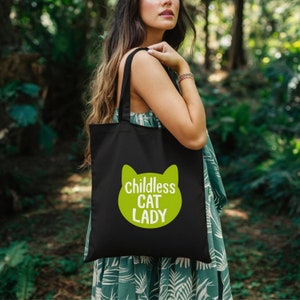 May include: Black tote bag with a lime green cat face design and the words "childless CAT LADY" in white. The bag is carried over the shoulder and is made of a sturdy fabric. The design is simple and eye-catching.