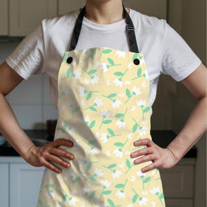 K&ouml;nnte beinhalten: Eine gelbe Sch&uuml;rze mit einem Blumenmuster aus wei&szlig;en Bl&uuml;ten und gr&uuml;nen Bl&auml;ttern. Die Sch&uuml;rze hat einen schwarzen Nackenriemen und schwarze Kn&ouml;pfe. Die Sch&uuml;rze wird &uuml;ber einem wei&szlig;en T-Shirt getragen.