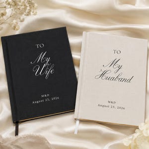 Può includere: Due libri con copertina rigida in tessuto. Il libro nero presenta la scritta bianca TO My Wife, M&D, August 23, 2026. Il libro color crema presenta la scritta nera TO My Husband, M&D, August 23, 2026. Entrambi hanno i bordi delle pagine dorati.