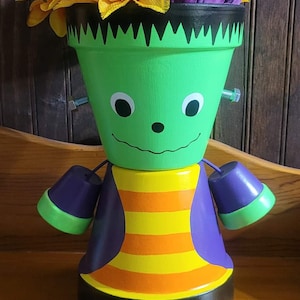 May include: A green and purple Frankenstein planter with yellow and purple flowers. The planter has a smiling face with white eyes and a black nose. The planter has green legs and black shoes.