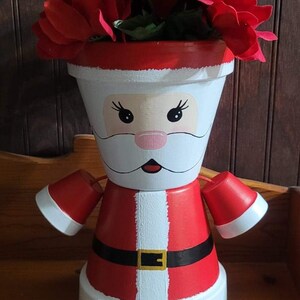 以下が含まれることがあります： 赤い帽子、白いひげ、黒いベルトでサンタクロースのようにペイントされた白いセラミックプランター。プランターには、赤いポインセチアの花束が入っています。