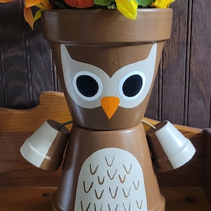 May include: A brown owl planter with white and brown accents. The planter is decorated with a white owl face and a brown beak. The planter is sitting on a wooden shelf and has orange flowers in it.