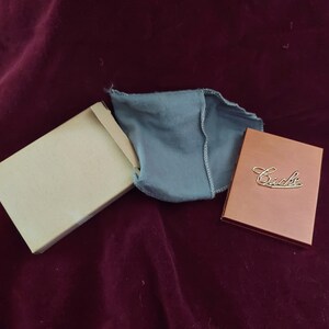 May include: A brown box with the word "Cacha" in gold lettering on the top. The box is sitting on a burgundy velvet surface. A blue velvet pouch is also on the surface.