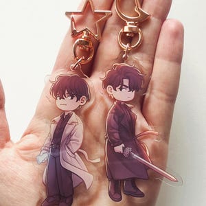 Pode incluir: Dois chaveiros de personagens de estilo anime com pingentes de estrela dourada e lua crescente. Os personagens são retratados em estilo chibi, um com um casaco branco e o outro com um casaco escuro com uma espada.