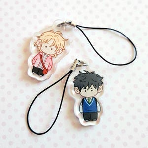 May include: Two clear acrylic charms featuring cartoon characters. One charm depicts a blonde character in a pink jacket with a red bag. The other charm shows a dark-haired character in a blue vest and white shirt. Both have black phone straps.