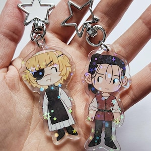 Könnte beinhalten: Zwei Schlüsselanhänger im Anime-Stil mit Zeichentrickfiguren. Ein Schlüsselanhänger hat eine blonde Figur mit Augenklappe, der andere eine Figur mit dunklem Haar. Beide Schlüsselanhänger haben sternförmige Verschlüsse und sind mit sternförmigem Glitzer verziert.