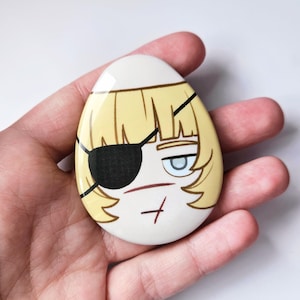 Puede incluir: Un pin con forma de huevo que presenta una ilustración de dibujos animados de un personaje con cabello rubio, un parche negro sobre un ojo y un solo ojo azul visible. El personaje tiene una tez pálida con una pequeña marca roja en la mejilla y la barbilla.