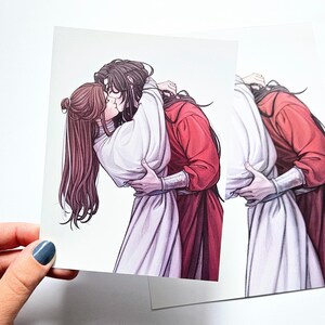 Puede incluir: Una impresión artística cuadrada con una ilustración digital de dos figuras de cabello largo y oscuro abrazándose y besándose. Una figura viste una túnica blanca y la otra una túnica roja con puños plateados. Estilo de líneas definidas.