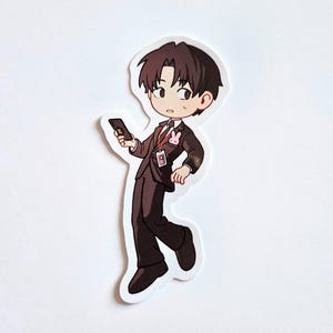 Puede incluir: Una pegatina troquelada que muestra un personaje de dibujos animados con cabello castaño oscuro, vestido con traje marrón oscuro, camisa blanca y corbata negra. El personaje sostiene un smartphone negro, lleva un cordón rojo y un pin de conejo rosa.