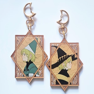 May include: Two rectangular acrylic keychains with gold-toned moon-shaped clasps. Each features a profile illustration of a blonde character in a geometric frame. One character wears a teal pointed hat, the other a black wide-brimmed witch hat.