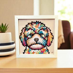 May include: A framed, colorful dog portrait. The artwork features a layered paper design with a white frame. The dog's fur is a mix of vibrant colors, including blue, orange, green, and pink. The background is a neutral beige.