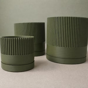 May include: Three cylindrical olive green planters of varying sizes, each featuring a vertical ribbed texture on the upper portion and a smooth, solid base. The planters are arranged in a cluster, showcasing their uniform matte finish and design.