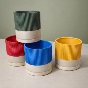 May include: A set of four cylindrical plant pots with vertical ribbed textures. Each pot features a two-tone design with a solid coloured upper section in dark green, red, blue, or yellow, and a contrasting light beige base and matching saucer.