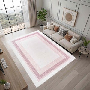 May include: A rectangular area rug with a white center and a pink border. The border features a gradient of pink shades, creating a layered effect. The rug is placed in a living room setting with a sofa and large window.