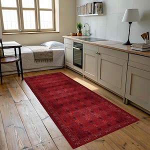 May include: A rectangular area rug featuring a deep red base colour with a repeating dark geometric pattern and a detailed border. The rug is positioned on a light-coloured wooden floor in a room with kitchen cabinetry and a bed visible in the background.