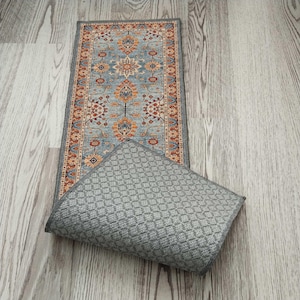 May include: A rectangular rug with a detailed floral pattern in shades of blue, orange, and red. The rug is bordered by a dark gray trim. The underside of the rug is a solid gray color with a diamond pattern.