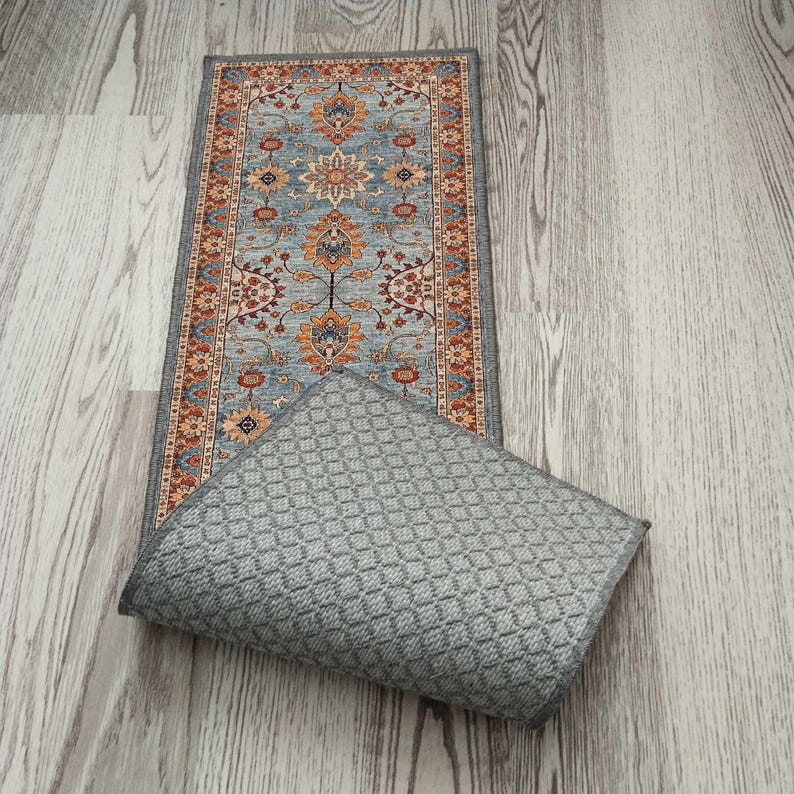 May include: A rectangular rug with a detailed floral pattern in shades of blue, orange, and red. The rug is bordered by a dark gray trim. The underside of the rug is a solid gray color with a diamond pattern.